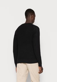 Suéter negro de punto con mangas largas, cuello redondo y dobladillo acanalado. La tela tiene una apariencia texturizada. Se lleva con pantalones beige.