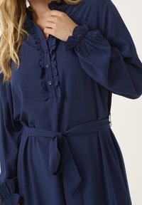 Abito blu navy con scollatura arricciata e pattina frontale, cinquecento in vita con maniche lunghe che presentano polsini arricciati. Tessuto con una finitura liscia.