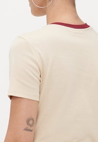 Beige t-shirt with a rounded neckline and contrasting maroon trim, short sleeves, and smooth fabric texture. Skin-tone arm features a sun tattoo.