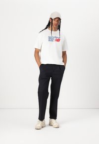 White cotton t-shirt with blue and red text, paired with dark navy trousers and beige sneakers. The cap is light gray.