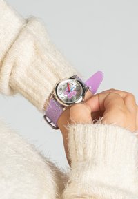 Montre en silicone violet avec paillettes, boîtier rond en argent, motif papillon sur le cadran et indicateurs numériques. Portée sur un pull blanc duveteux.