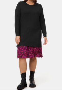 Robe en maille noire à manches longues avec un col rond, superposée à une jupe rose à motif floral, associée à des bottines noires.