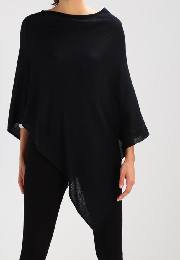 Femme portant un poncho asymétrique noir sur un pantalon noir, debout devant un fond gris clair uni.