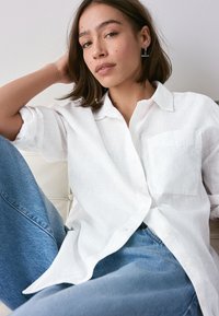Weißes, strukturiertes Hemd mit Knopfleiste und Brusttasche, kombiniert mit hellblauen Jeans. Modell ruht auf einer weißen Oberfläche.