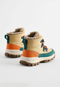 Bottes d'hiver colorées avec une tige kaki, une doublure en fausse fourrure, des accents orange et bleu sarcelle, et une semelle robuste avec des rainures profondes pour l'adhérence.