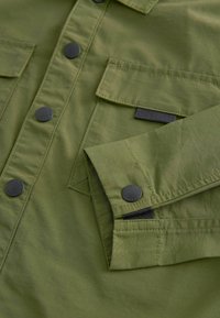 Giacca camicia verde oliva con bottoni a pressione neri, due tasche sul petto e un tessuto texturizzato. Include un'etichetta con un accento nero.
