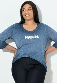 Niebieska koszulka z dekoltem w serek i rękawami do połowy przedramienia, z białym napisem "MOIN", ozdobiona motywem kotwicy i butelki. Ciemne dżinsy.