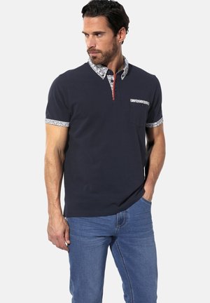 Navy polo shirt with floral-patterned collar and sleeves. Features a chest pocket and a red button placket. Paired with blue jeans.