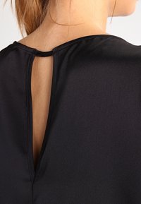 Black top with a keyhole back detail. Smooth fabric, subtle sheen, and a round neckline. Seam and stitch detailing along the edges.