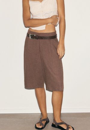 Femme portant un short large marron mi-genou avec une ceinture noire, un haut blanc sans manches, et des sandales marron à brides croisées, debout contre un mur uni.