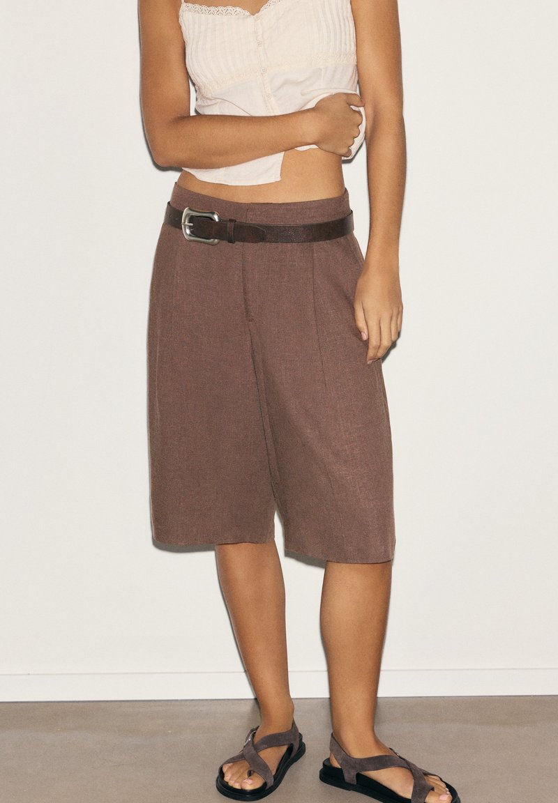 Femme portant un short large marron mi-genou avec une ceinture noire, un haut blanc sans manches, et des sandales marron à brides croisées, debout contre un mur uni.