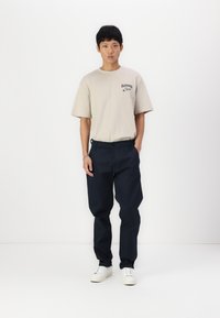 Bež majica iz bombaža kratkih rukava s prednjim printom, uparena s tamnoplavim hlačama i bijelim tenisicama, stoji na jednobojnoj pozadini.