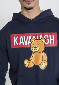 Sudadera con capucha en azul marino que presenta un gran gráfico de un oso de peluche amarillo con una sonrisa tachada. El texto dice "KAVANAGH" en letras rojas en negrita.