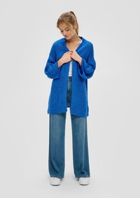 Blauwe oversized gebreide cardigan met brede mouwen, open voorkant en textuurstof; gecombineerd met lichte denim wijde broek en witte sneakers.