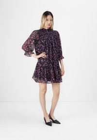 Kvinne iført en gjennomsiktig, mørk blomsterkjole med puffermer og svarte spisse slingback-hæler, stående med den ene hånden på hoften.