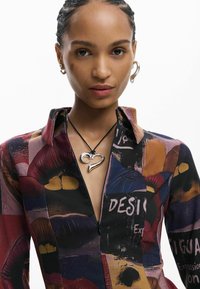 Cămașă cu mâneci lungi, cu imprimeu grafic colorat cu buze pe un fundal negru. Este vizibil un pandantiv argintiu în formă de inimă pe un șnur negru.