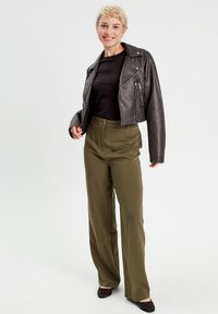 Veste en cuir noir coupée de motard, haut noir ajusté, pantalon vert olive taille haute et ballerines noires, sur fond blanc uni.