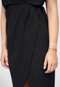 Black dress with a textured fabric, featuring a twisted waist detail, and a side slit. The hand shown wears a simple silver bracelet.