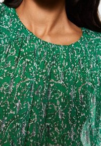 Haut vert avec un col froncé, motifs floraux et de vigne blancs complexes, et accents métalliques argentés. Tissu léger avec texture.