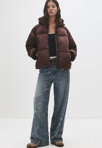 Braune Puffjacke mit Kapuze, im kastigen Design und mit steppmuster. Kombiniert mit weiten, distressed Jeans.