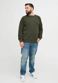 Pull en tricot vert foncé avec un col rond, associé à un jean en denim clair délavé et des baskets blanches avec des rayures noires.