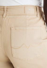 Béžové džíny s texturovaným povrchem, s zadní kapsou se šitým vlnkovým vzorem a značkovou nášivkou na pásku.