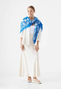 Écharpe à motif floral bleu drapée sur un blouse blanche et une longue jupe crème, associée à des tongs blanches à plateforme.