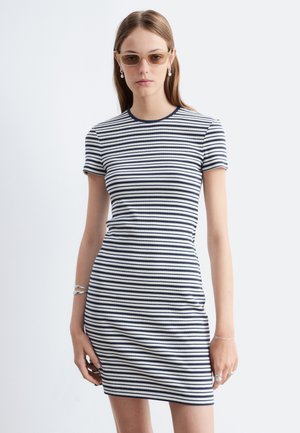 Femme portant des lunettes de soleil beige et une robe à manches courtes rayée bleu marine et blanc, debout devant un fond clair uni.
