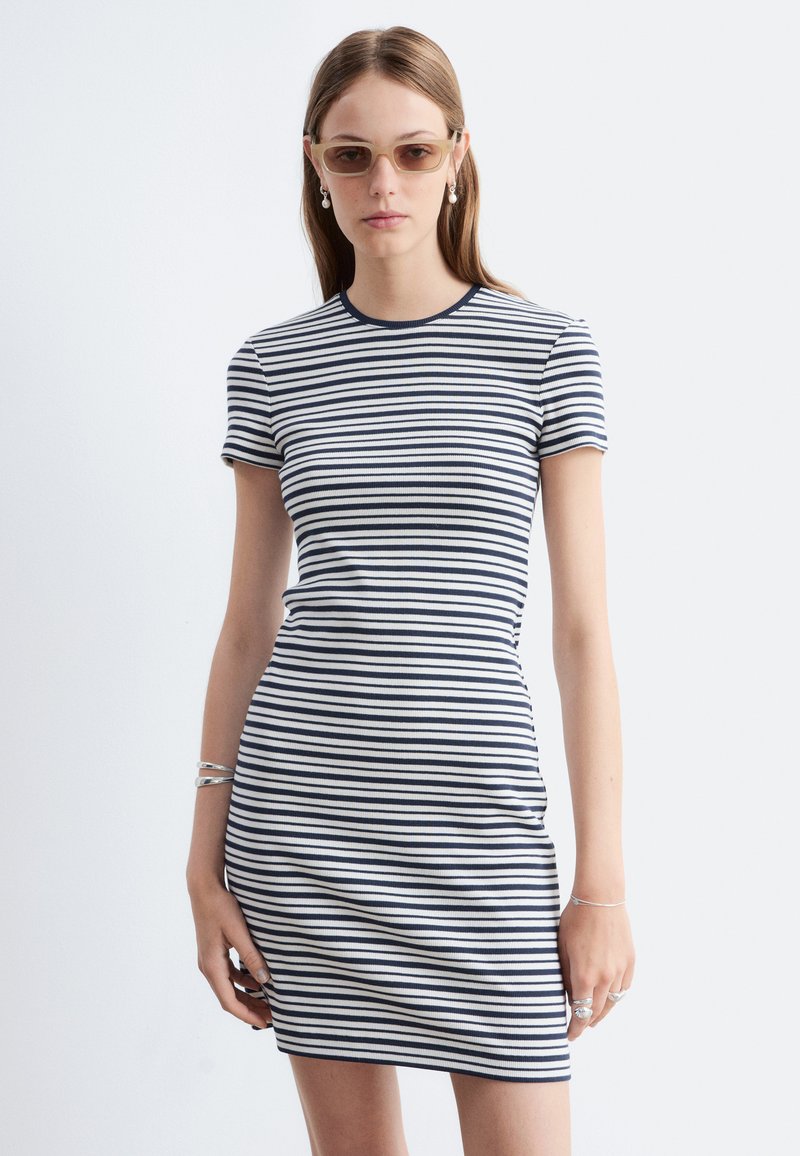 Femme portant des lunettes de soleil beige et une robe à manches courtes rayée bleu marine et blanc, debout devant un fond clair uni.
