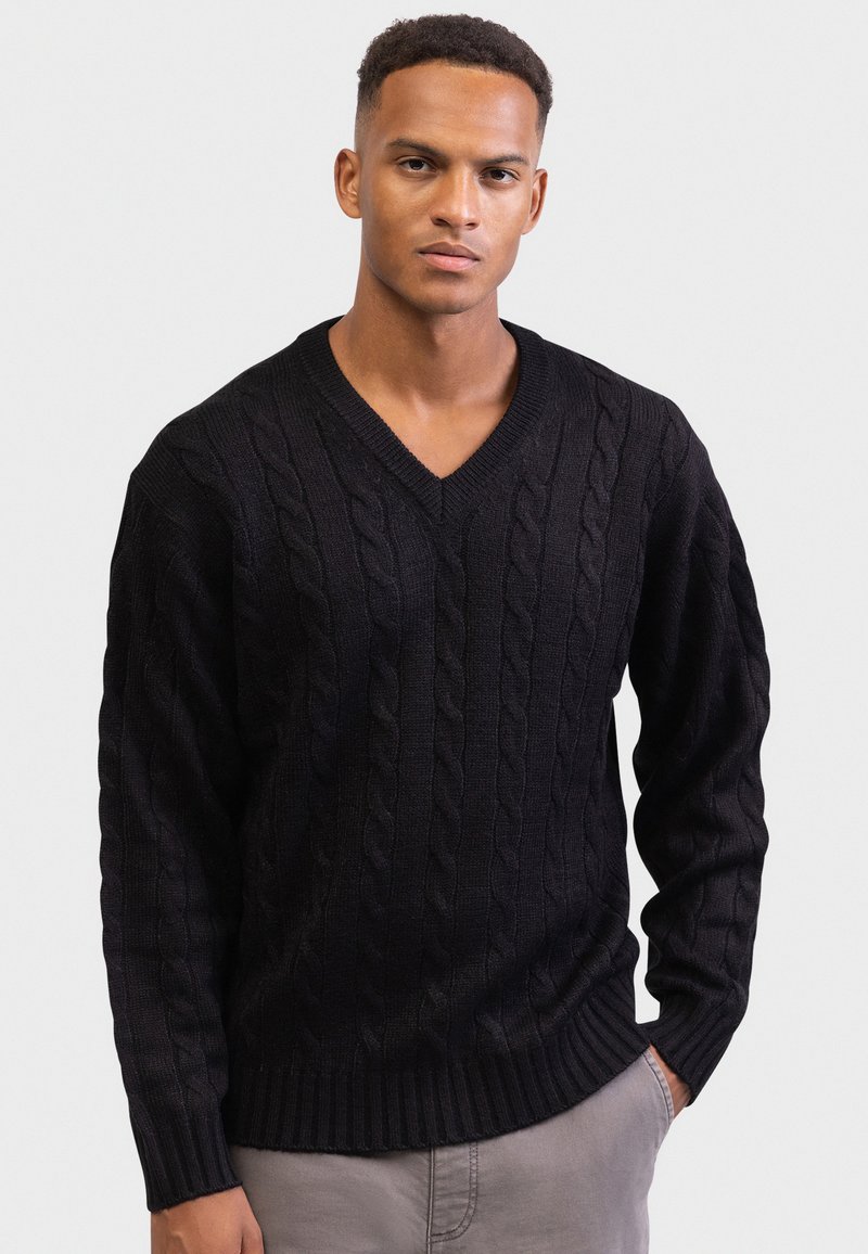 Czarny sweter w warkoczowy splot z dekoltem w kształcie litery V, teksturowanym wzorem, ściągaczami na rękawach i u dołu. Noszony na szarych spodniach na jasnym tle.