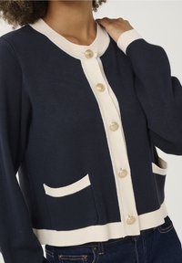 Navyfarbener Cardigan mit elfenbeinfarbenem Besatz, ausgestattet mit goldenen Knöpfen und zwei Fronttaschen. Der Stoff wirkt strukturiert und weich.