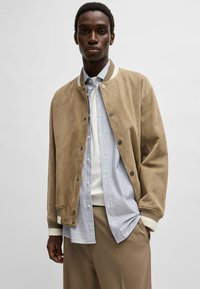 Tan suède bomberjack met ribbelboorden en kraag, met drukknoopsluitingen, gelaagd over een gestreepte shirt, gedragen met beige broeken.