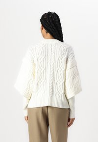 Maglione bianco lavorato a maglia caratterizzato da un motivo a treccia spesso, maniche oversize e polsini a costine. Indossato con pantaloni beige.