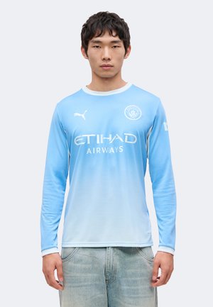 Młody mężczyzna noszący jasnoniebieską, długorękawową koszulkę piłkarską Manchester City z logo Etihad Airways i Puma, w połączeniu z jasnymi dżinsami.