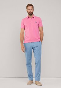 Polo shirt da uomo rosa con maniche corte, caratterizzato da un colletto e un piccolo logo. Abbinato a pantaloni azzurri chiari e scarpe beige senza lacci.