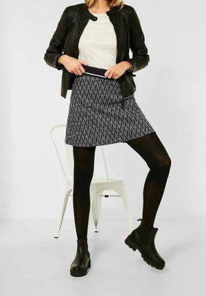 Schwarze Lederjacke über einem weißen Oberteil, kombiniert mit einem gemusterten schwarz-weißen Miniröckchen, schwarzen Strumpfhosen und klobigen schwarzen Stiefeln.