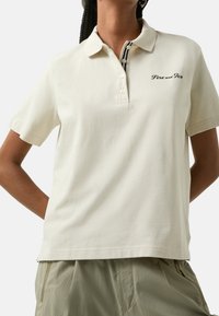 Polo de couleur crème en tissu doux, avec un col, trois boutons, et un logo brodé « Fire and Ice » sur la poitrine.