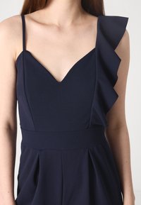 Ναυτικό μπλε φόρεμα με V-λαιμό, έναν ώμο με ρυτίδωση, εφαρμοστό κορμό και φαρδιά παντελόνια σε πλισέ σχέδιο από μαλακό ύφασμα.