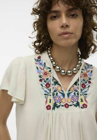 Lys beige bluse med korte, puffede ærmer, der har farverig blomsterbroderi omkring halsudskæringen og plisseret tekstur. Sølv perle halskæde.
