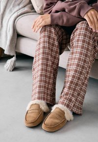 Chaussons en suède marron avec doublure en fourrure, associés à un pantalon de pyjama à carreaux beiges, marron et rouille.