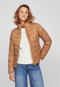 Kohvipruun puffer-jakk tan, horisontaalsete õmblustega, kõrge kaelusega ja lukuga. Kanda kreemivärvi palmikseeliku sviitri ja siniste teksade peal.