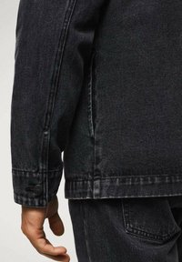 Chaqueta de mezclilla negra con una silueta estructurada, costuras visibles y un detalle de botón en el puño. La mano descansa a un lado.