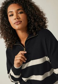Schwarzer Zip-Up-Pullover mit breiten cremefarbenen Streifen, ausgestattet mit einem goldenen Reißverschluss und einer weichen Textur. Das Design ist lässig mit einem hohen Kragen.