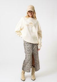 Sweatshirt crem cu logo-ul "HARMONY", asortat cu o fustă din material leopard cu o fanta laterală și cizme cowboy bej. Pe cap poartă o șapcă bej.