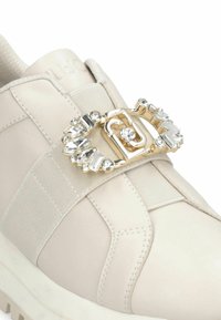 Sneaker en cuir crème avec sangle élastique et boucle en tonalité dorée ornée de cristaux décoratifs transparents.