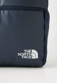 Dunkelblauer Rucksack mit einem Karomuster, ausgestattet mit einem weißen Logo, einem horizontalen Reißverschluss und strukturiertem Stoff. Einfaches, rechteckiges Design.