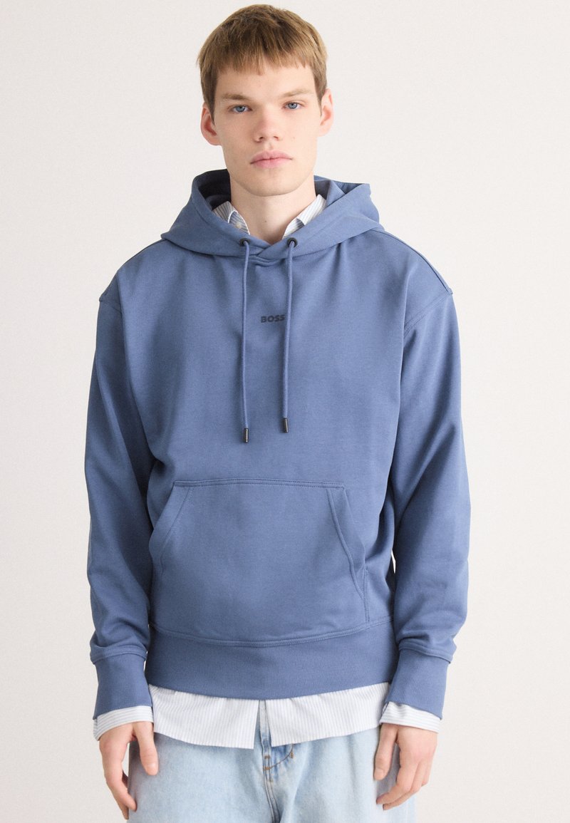 Giovane uomo che indossa un hoodie blu con il logo "BOSS", sopra una maglietta a righe, e jeans azzurri chiari, in piedi su uno sfondo semplice.