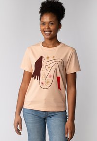 Pfirsichfarbene Baumwoll-T-Shirt mit Rundhalsausschnitt, das eine Grafik einer in Braun umrandeten Figur zeigt, die mit floralen Details und roten Akzenten geschmückt ist.