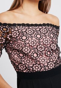 Haut en dentelle noire à épaules dénudées avec un motif floral sur une couche inférieure rose, doté d'un design texturé et d'un corsage noir ajusté.