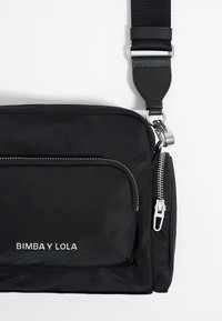 Borsa a tracolla nera in nylon con hardware argentato, dotata di una tasca frontale con zip, dettagli con logo e tracolla regolabile. Texture liscia e forma rettangolare.